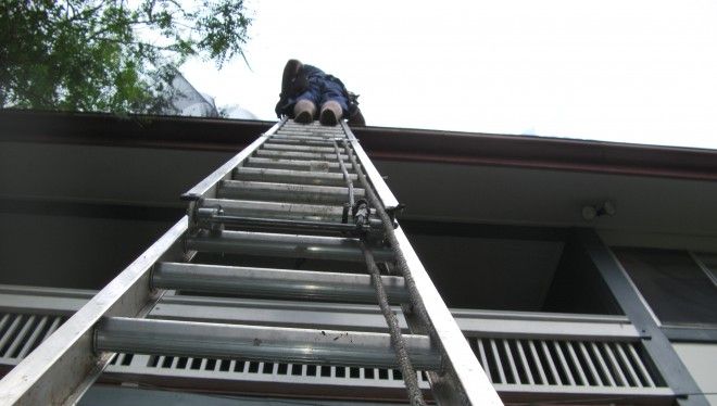 looking up at a man on a ladder installing gutter mesh onto a roof.